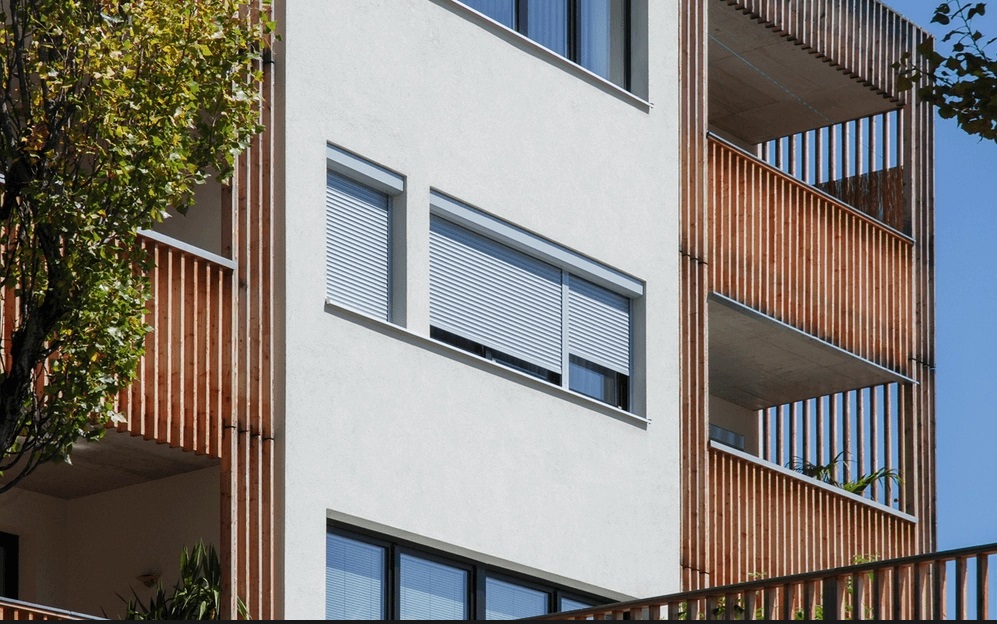 Moderní bytový dům s předokenními roletami v bílé barvě; elegantní kombinace s dřevěným lamelovým obkladem lodžií pro účinné stínění a soukromí.